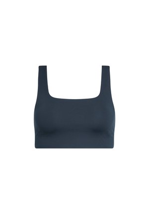 Tumejuustega naine hobusesabaga, kes kannab tumerohelist lühikest topi, musti vabaaja pükse ja halli spordijalatsit, seisab ühe käega taskus.