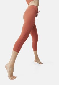 Capri tights i rustfarge, laget av elastisk stoff. Har høy midje med snøring og sømdetaljer langs bena.