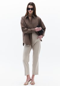 Hnědá košile s knoflíky, béžové přizpůsobené kalhoty a zlaté sandály na podpatku. Modelka drží černou kabelku. Jednoduché bílé pozadí.