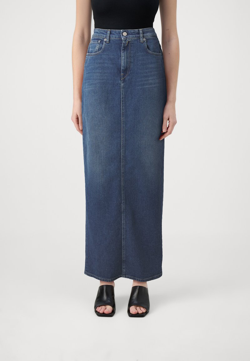 Jupe maxi en denim bleu foncé, avec une taille haute, coupe droite et un léger dégradé. Portée avec des sandales à talons ouverts noires.