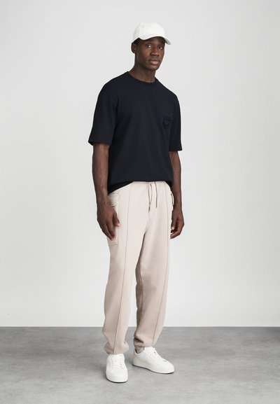 Svart kortermet t-skjorte med brystlomme, beige joggebukser med sidelommer, og hvite sneakers, kombinert med en hvit caps.