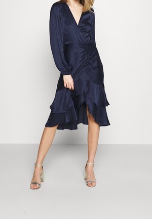Marineblå satinkjole med lange ærmer, V-udskæring, rynkede detaljer og lagdelt flæsekant. Kombineret med metallic hælede sandaler.