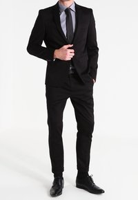 Mann in einem schwarzen Anzug, hellblauem Hemd, schwarz-weißer gepunkteter Krawatte und schwarzen Lederschuhen, steht mit einer Hand in der Tasche.