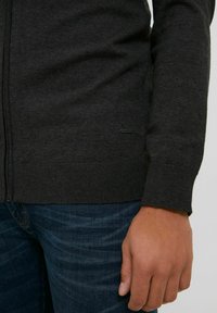 Mörkgrå zip-up kofta i stickad tyg, med ribbade ärmslut och nederkant, samt en liten varumärkesetikett längst ner till vänster. Bärs ihop med blå jeans.