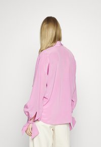 Blusa di raso rosa con maniche lunghe, con dettagli a fiocco ai polsini e un colletto semplice. Texture liscia, vestibilità comoda.