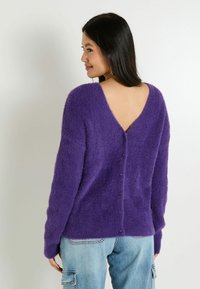 Suéter morado esponjoso con un diseño de espalda en V profunda, con detalles de botones a lo largo de la columna vertebral. Mangas largas y textura suave.
