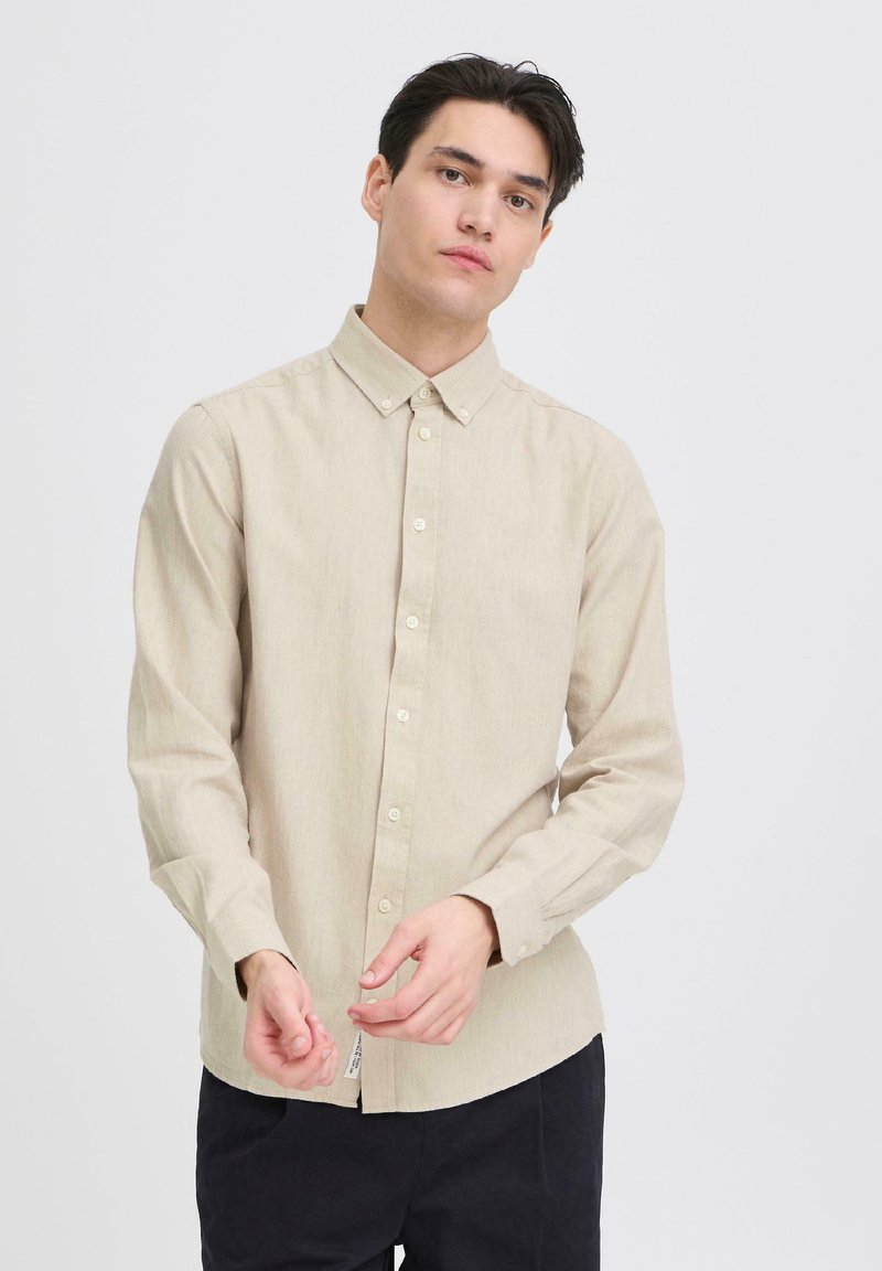 Chemise beige à manches longues avec un col boutonné, texture unie et fermeture à boutons sur le devant. Le modèle ajuste les poignets, porte un pantalon foncé.