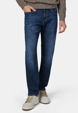 Mann trägt dunkelblaue Straight-Leg-Jeans und beige Freizeitschuhe, mit einer Hand in der Tasche und einem grauen Pullover, der an der Taille sichtbar ist.