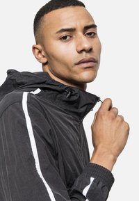 Chaqueta ligera negra con capucha, con franjas blancas a los lados, cierre de cremallera y puños elásticos. Tejido texturizado, diseño deportivo.