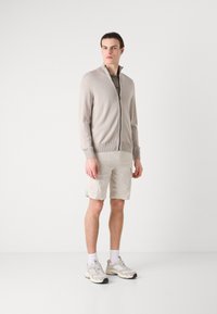 Lys beige glidelås cardigan laget av strikket materiale, kombinert med lyse shorts og grå joggesko. Rent design med ribbede mansjetter og kant.
