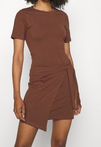 Robe brune à manches courtes, fabriquée en tissu doux avec une coupe ajustée, dotée d'un détail de lien enroulé à la taille et d'un ourlet asymétrique.