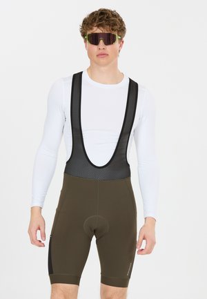 Junger Mann mit reflektierenden Sportsonnenbrillen, weißem Langarmshirt und olivgrünen Radhose mit Netzträgern.