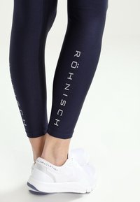 Navyblå leggings med blank yta, med vertikal "RÖHNISCH"-logotyp i vitt på sidan. Bärs med vita träningsskor.