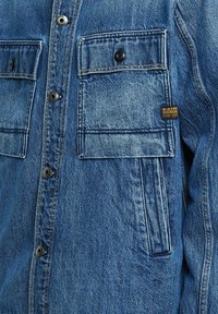 Giacca in denim blu vista frontalmente, con due tasche sul petto con bottoni e tasca laterale con fessura, cuciture visibili e etichetta G-Star Raw.