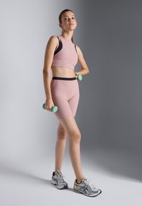 Sportoutfit met een geribbelde roze crop top en bijpassende shorts, zwarte tailleband, grijze sneakers en houdt lichtgroene dumbbells vast.