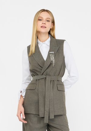 Gilet grigio a spina di pesce con vita cinturata, revers a sprone e due tasche frontali. Indossato sopra una camicia bianca con maniche a volant.