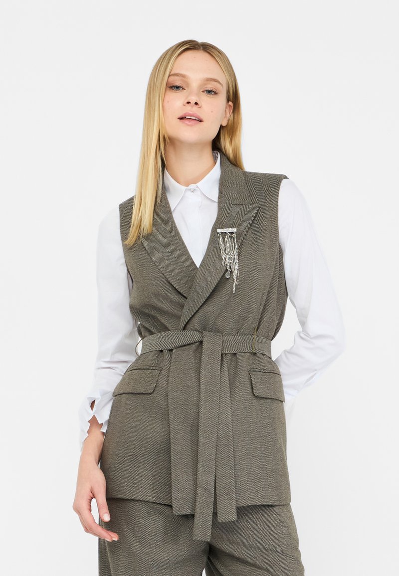 Gilet grigio a spina di pesce con vita cinturata, revers a sprone e due tasche frontali. Indossato sopra una camicia bianca con maniche a volant.