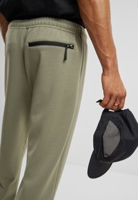 Jogging en vert olive clair avec une poche zippée à l'arrière, associé à une casquette noire avec un panneau en maille. Texture du tissu lisse visible.