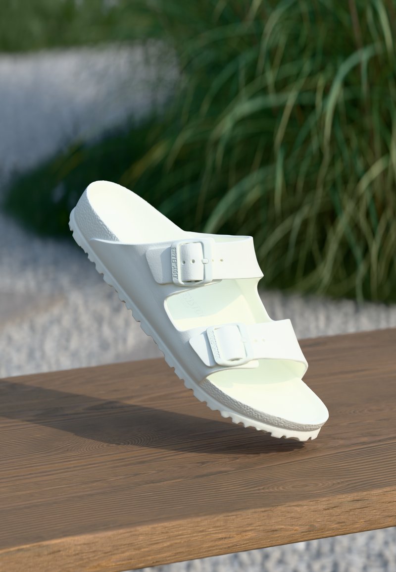 White sandals with two adjustable buckles, made of smooth synthetic material. Thick, textured sole with a slight curve and rounded shape.
