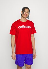Sarkana kokvilnas T-krekls ar baltu "adidas" logo uz krūtīm. Ar īsām piedurknēm un apaļu kakla izgriezumu. Valkāts ar ziliem šortiem.