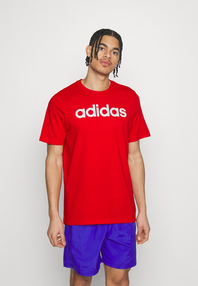 Sarkana kokvilnas T-krekls ar baltu "adidas" logo uz krūtīm. Ar īsām piedurknēm un apaļu kakla izgriezumu. Valkāts ar ziliem šortiem.