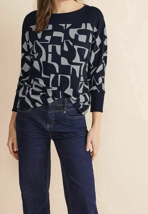 Femme portant un haut à manches longues bleu marine et gris à motif abstrait et un jean en denim bleu foncé, debout devant un fond beige.