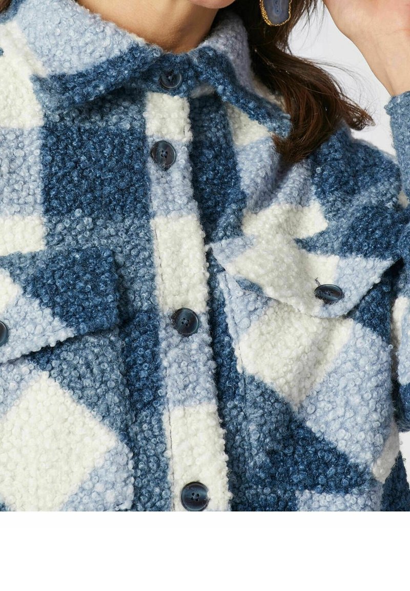 Donna che indossa un cappotto di lana a quadri blu e bianchi con grandi bottoni e tasche a patta, mentre tiene il suo orecchino vicino all'orecchio.