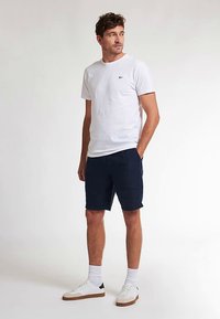 Valkoinen lyhythihainen t-paita, jossa pieni musta logo, yhdistettynä tummansinisiin shortseihin ja valkoisiin lenkkareihin, joissa on vaaleanruskeat yksityiskohdat. Malli seisoo rennosti.