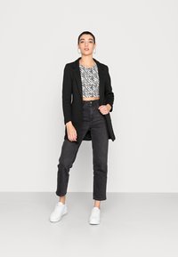 Blazer nero lungo su un top a strisce zebrate corto, abbinato a jeans neri a vita alta e sneakers bianche. Design semplice e strutturato.