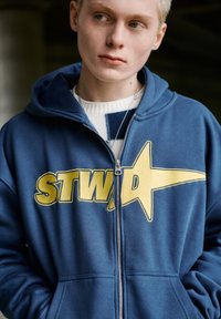 Young person with short blonde hair wearing a blue zip-up hoodie labeled "STWA" over a white and navy sweater.