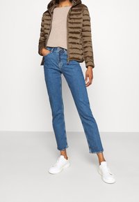 Bruine donzen jas met capuchon, een beige top, blauwe recht gesneden jeans en witte sneakers tegen een neutrale achtergrond.