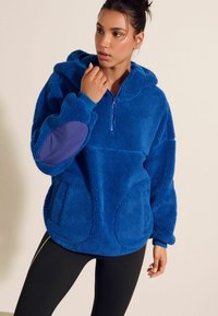 Blå fleece-pullover med dragkedja i halsen, en huva och långa ärmar. Har matchande blå stretchmuddar och en lila armbågsplatta.