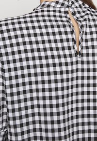 Czarna i biała koszula w kratkę typu gingham z wiązanym wycięciem w kształcie klucza z tyłu oraz krótkimi rękawami. Materiał jest lekki i w kratkę.