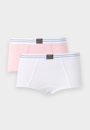 Jeune femme debout pieds nus dans un ensemble de sous-vêtements assortis rose clair avec des bandes élastiques blanches et un logo noir sur fond blanc uni.