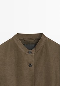 Kraag en bovenkant voorkant van een bruin linnen overhemd met twee zwarte knopen en een mandarin kraag op een witte achtergrond.