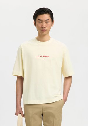 Junger Mann trägt ein übergroßes cremefarbenes T-Shirt mit rotem Schriftzug "HÔTEL AMOUR", beigefarbene Hose, hält eine beigen Einkaufstasche, neutraler Hintergrund.