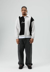 Wit-zwarte pullover met ritssluiting en kraag, met een geborduurd logo, gecombineerd met losse zwarte jeans en zwarte sneakers.