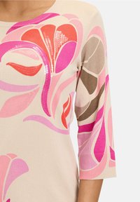 Light beige knit top featuring vibrant pink, red, and brown floral patterns with sequin accents. Three-quarter sleeves and ribbed neckline.