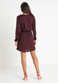 Una mujer con cabello largo y marrón, vestida con un vestido burdeos de manga larga y sandalias de tacón coral, de pie de espaldas contra un fondo blanco.