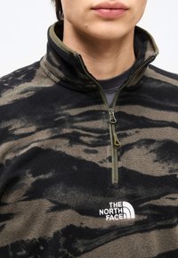 Άτομο που φοράει φλις με μαύρο και ελιά μοτίβο, με φερμουάρ μέχρι το στήθος και το λογότυπο "The North Face" στο στήθος.