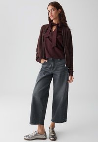 Bordeaux blouse met strikdetail, bruine fluffy cardigan, grijze wijde jeans en metallic sneakers. Textuurstoffen en een comfortabele pasvorm.