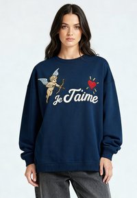 Femme portant un sweat-shirt bleu marine avec un cupidon, un cœur rouge et le texte "Je T’aime", associé à un jean délavé foncé.