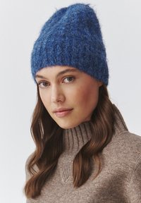 Caciulă tricotată albastră, cu o textură pufoasă, design conic și margine striată. Purtată cu un pulover turtleneck în nuanță maro deschis, cu striații.