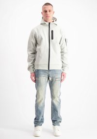 Lichtgrijze zip-up hoodie met zwarte rits en zakaccenten; gedragen met versleten blauwe spijkerbroek en witte sneakers.