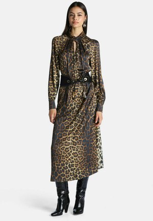 Femme portant une longue robe à imprimé léopard avec un col noué, ceinture large noire, bottines noires à talons et boucles d'oreilles remarquables, debout devant un fond blanc.