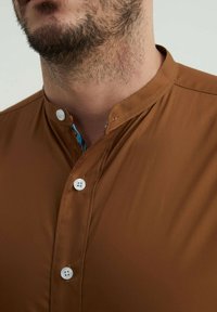 Chemise marron à col mandarin, dotée de boutons blancs et d'un col intérieur à motifs colorés. Texture de tissu lisse.