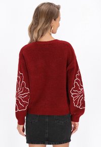 Červený pletený svetr s dlouhými rukávy, který má bílé květinové vyšívání a perlové detaily. Měkká textura, volný střih a žebrovaný lem.
