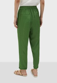 Person wearing green patterned tapered pants with elastic waistband and beige flat sandals, standing on gray floor.