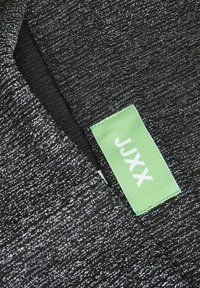 Sort stof med metallisk sølvtråd, der har et grønt mærke med hvid tekst, der står "JJKX." Teksturen ser blød og strikket ud.