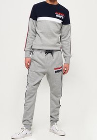 Grijze sweatshirt met marineblauwe en witte strepen, voorzien van het "Superdry" logo. Grijze joggers met trekkoord, zijaccenten en bijpassend logo.
